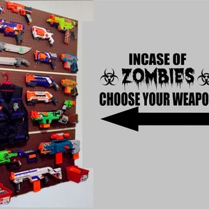 May include: A wall with a pegboard displaying a collection of toy guns, a black tactical vest, and a sign that reads "In case of zombies choose your weapon".