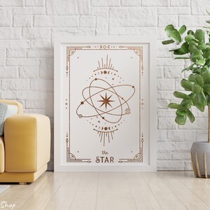 May include: A white framed print with a gold geometric design of a star with a circle around it. The text "The STAR" is at the bottom of the print. The print is hanging on a white brick wall with a green plant in a gray pot to the right.