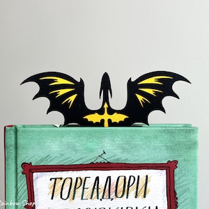 May include: A black and yellow dragon-shaped bookmark sits atop a book. The bookmark features a dragon silhouette with yellow wing details. The book has a teal cover with text in a red framed box.
