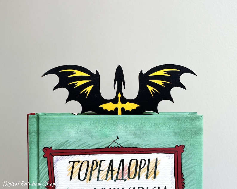 May include: A black and yellow dragon-shaped bookmark sits atop a book. The bookmark features a dragon silhouette with yellow wing details. The book has a teal cover with text in a red framed box.
