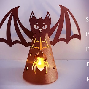 May include: A brown paper bat lantern with a spider and web design. The bat has large wings and a cute face. The lantern is lit from inside, creating a warm glow.