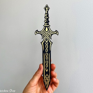 May include: Black and gold decorative sword with intricate detailing. The sword features a stylized design with a gold handle and blade accents. The sword is held against a light gray background.