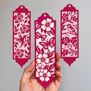 Puede incluir: Tres marcapáginas decorativos magenta con intrincados diseños recortados. Uno muestra un colibrí, otro mariposas y el tercero un pájaro en una rama. Cada marcapáginas tiene un patrón floral único y una base decorativa.