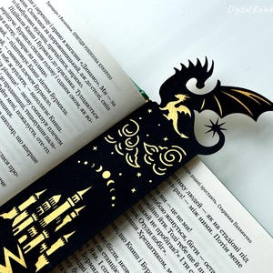 May include: A black and gold dragon bookmark with a castle design. The bookmark features a dragon silhouette with outstretched wings, a star, and a castle illustration. The bookmark is inserted into a book with text.