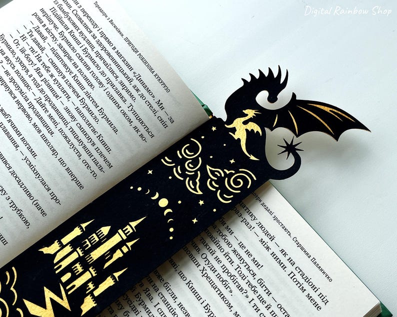 May include: A black and gold dragon bookmark with a castle design. The bookmark features a dragon silhouette with outstretched wings, a star, and a castle illustration. The bookmark is inserted into a book with text.
