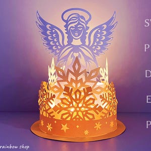 Puede incluir: Un portavelas de papel con un delicado ángel con alas y un diseño de copo de nieve. El portavelas está iluminado desde el interior, creando un brillo cálido.