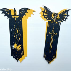 May include: Two fantasy-themed bookmarks with dragon and sword designs. The bookmarks are black and yellow, with intricate cut-out details. One features a dragon with a sword, the other a dragon with a book and flames. Ideal for book lovers.