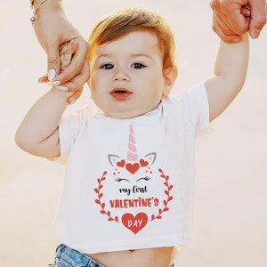 Puede incluir: Una camiseta blanca con un gr&aacute;fico de unicornio rosa y el texto "my first valentine's day" en rojo. El unicornio tiene corazones rosas y rojos alrededor de su cabeza.