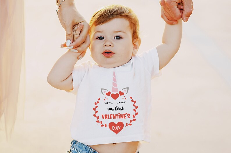 Puede incluir: Una camiseta blanca con un gr&aacute;fico de unicornio rosa y el texto "my first valentine's day" en rojo. El unicornio tiene corazones rosas y rojos alrededor de su cabeza.