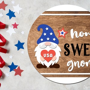 Puede incluir: Un letrero redondo de madera con un gnomo rojo, blanco y azul que sostiene un coraz&oacute;n con "USA" escrito en &eacute;l. El letrero dice "home sweet gnome" en letras blancas y negras.