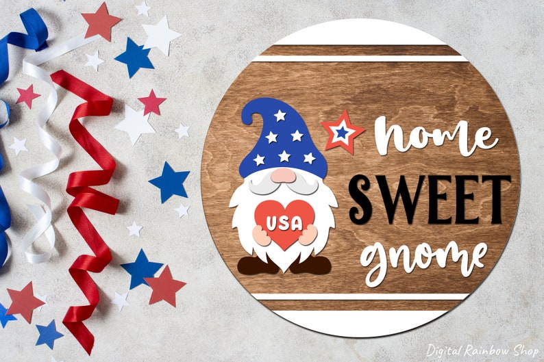 Puede incluir: Un letrero redondo de madera con un gnomo rojo, blanco y azul que sostiene un coraz&oacute;n con "USA" escrito en &eacute;l. El letrero dice "home sweet gnome" en letras blancas y negras.