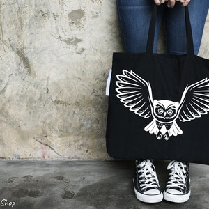 Op de afbeelding: Een zwarte tote bag met een witte uil afbeelding. De uil heeft grote, ronde ogen en uitgespreide vleugels. De tas wordt vastgehouden door iemand die een blauwe jeans draagt.
