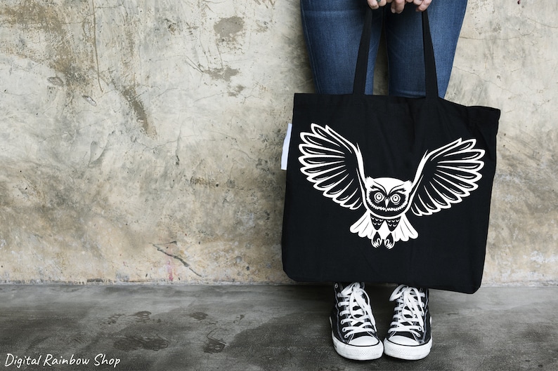 Op de afbeelding: Een zwarte tote bag met een witte uil afbeelding. De uil heeft grote, ronde ogen en uitgespreide vleugels. De tas wordt vastgehouden door iemand die een blauwe jeans draagt.