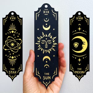 May include: Three black and gold tarot card bookmarks. The bookmarks feature celestial designs including the sun, moon, and stars. Each bookmark has a different design and the words "The Star", "The Sun", and "The Moon".