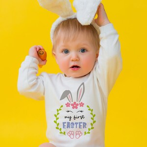 Puede incluir: Un beb&eacute; con un mono blanco con un gr&aacute;fico de conejo y el texto "my first easter" en una corona. El beb&eacute; lleva una diadema de orejas de conejo blancas y sostiene una zanahoria.