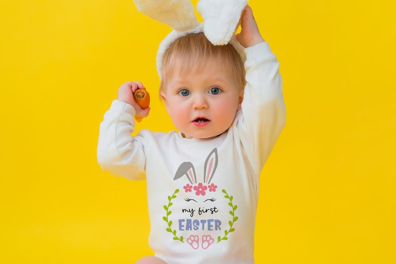 Puede incluir: Un beb&eacute; con un mono blanco con un gr&aacute;fico de conejo y el texto "my first easter" en una corona. El beb&eacute; lleva una diadema de orejas de conejo blancas y sostiene una zanahoria.