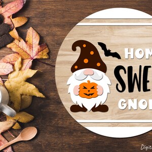 Puede incluir: Un letrero redondo de madera con un borde blanco y un fondo de madera marr&oacute;n. El letrero presenta un gnomo de dibujos animados sosteniendo una calabaza con una cara espeluznante. El texto "Home Sweet Gnome" est&aacute; escrito en letras blancas y negras.