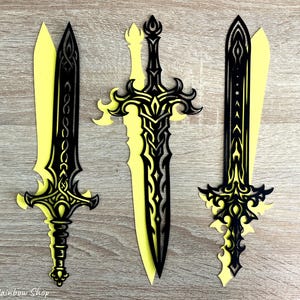 May include: Three decorative paper swords with intricate black designs layered over yellow. The swords have ornate handles and blades, with the middle sword being the largest. The background is a light brown wooden surface.