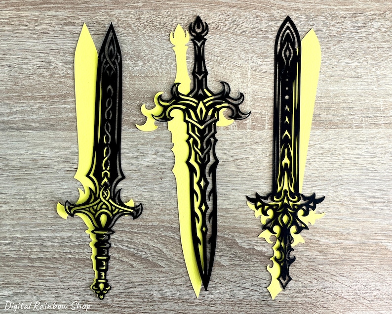 May include: Three decorative paper swords with intricate black designs layered over yellow. The swords have ornate handles and blades, with the middle sword being the largest. The background is a light brown wooden surface.