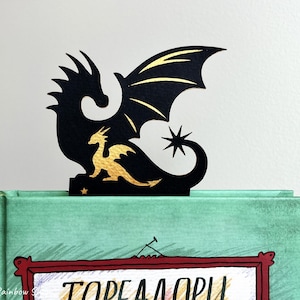 May include: A black dragon bookmark with gold detailing, placed on a green book. The bookmark depicts a large dragon silhouette with a smaller gold dragon and a star. The book has a red frame with text.