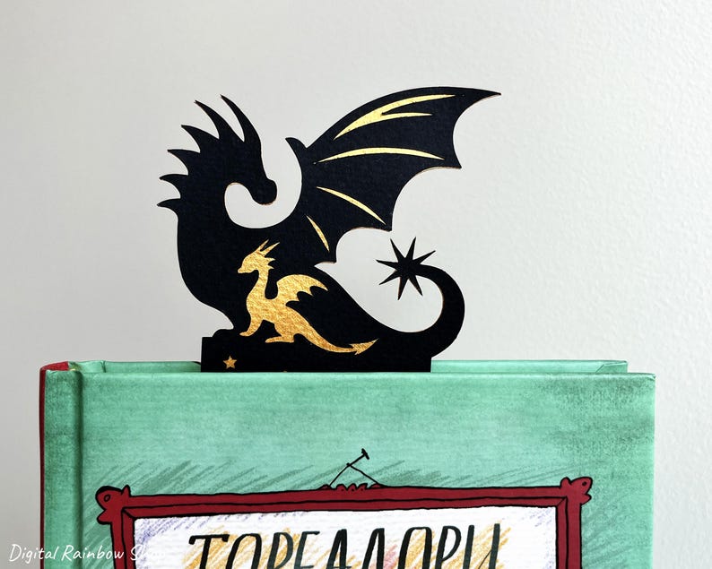 May include: A black dragon bookmark with gold detailing, placed on a green book. The bookmark depicts a large dragon silhouette with a smaller gold dragon and a star. The book has a red frame with text.