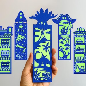 Puede incluir: Una mano sosteniendo cinco marcapáginas de papel azul y verde. Cada marcapáginas presenta un diseño diferente: un tren, un cohete, un dinosaurio, un pirata y un coche de policía. Los marcapáginas están recortados con detalles intrincados.
