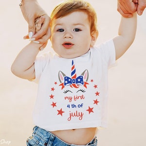 Puede incluir: Una camiseta blanca con un dise&ntilde;o de unicornio rojo, blanco y azul. El unicornio tiene un lazo con estrellas y el texto "my first 4th of july".