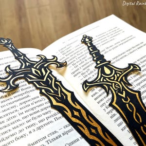May include: Two black and gold sword-shaped bookmarks rest on the pages of a book. The bookmarks have intricate designs and are made of a dark material with gold accents. The book is open, with text visible.