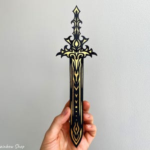 May include: A black and gold decorative sword-shaped bookmark. The bookmark features intricate gold filigree designs on a black background. The sword has a detailed hilt and blade, with a pointed tip. The bookmark is held in a hand.