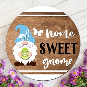 Puede incluir: Un letrero redondo de madera con un borde blanco y un gnomo sosteniendo un huevo de Pascua. El letrero dice "home sweet gnome" en letras blancas y negras.