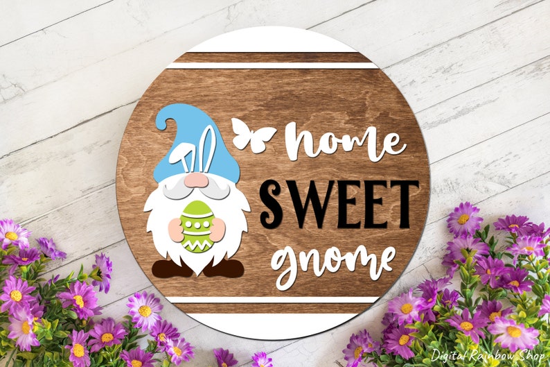 Puede incluir: Un letrero redondo de madera con un borde blanco y un gnomo sosteniendo un huevo de Pascua. El letrero dice "home sweet gnome" en letras blancas y negras.