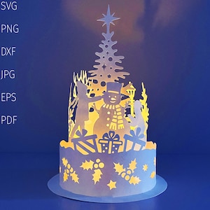 Puede incluir: Decoración navideña de papel en capas con un muñeco de nieve, un árbol de Navidad y regalos. El diseño está iluminado desde dentro, creando un brillo cálido. La base está decorada con recortes de acebo y bayas.