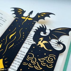 May include: Black dragon bookmarks with gold detailing, showcasing dragon silhouettes, stars, and cloud patterns. The bookmarks are positioned within a book, enhancing the fantasy theme. The bookmarks are made of a dark material.