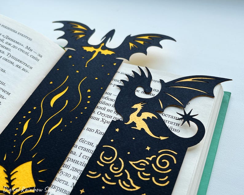 May include: Black dragon bookmarks with gold detailing, showcasing dragon silhouettes, stars, and cloud patterns. The bookmarks are positioned within a book, enhancing the fantasy theme. The bookmarks are made of a dark material.