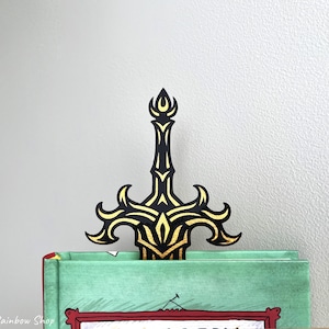 May include: A black and gold sword-shaped bookmark is inserted into a book. The bookmark has an ornate design with gold accents. The book has a green cover and red trim. The background is a plain, light gray wall.