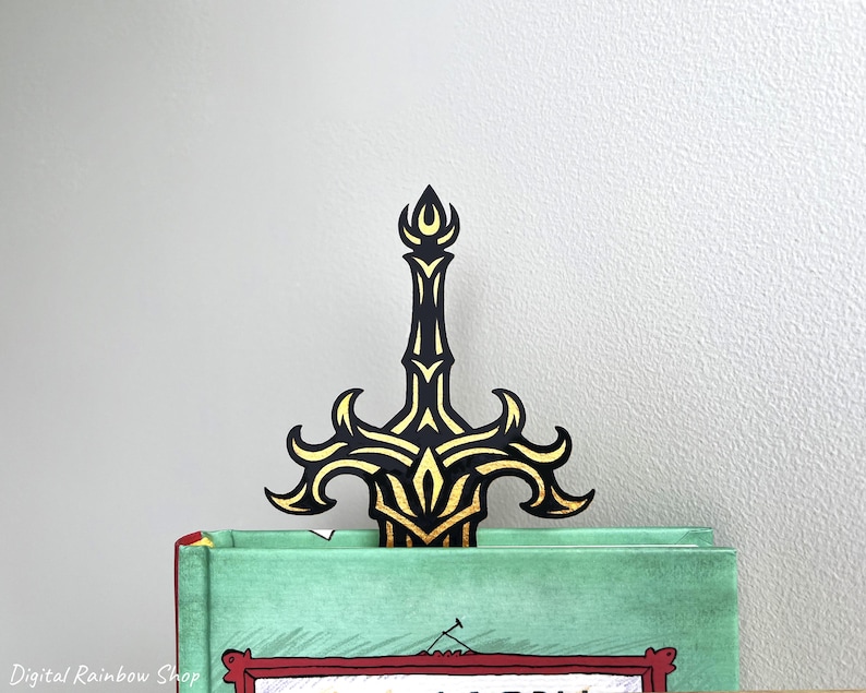 May include: A black and gold sword-shaped bookmark is inserted into a book. The bookmark has an ornate design with gold accents. The book has a green cover and red trim. The background is a plain, light gray wall.