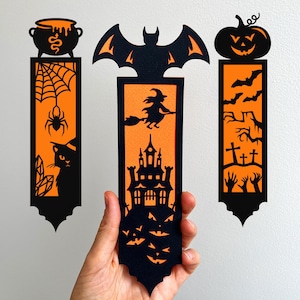 May include: Three Halloween-themed bookmarks in black and orange. One features a cauldron, spiderweb, and cat. Another shows a bat, witch, and castle. The last has a pumpkin, bats, and graveyard elements. The bookmarks are held by a hand.