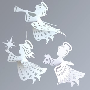 以下が含まれることがあります： 白の切り絵の天使のオーナメント3つ。1つはトランペットを吹き、もう1つは星を持ち、3つ目は贈り物を持っています。天使たちはグレーの背景に吊り下げられており、休日の装飾に最適です。