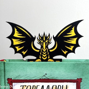 May include: A black and yellow dragon bookmark with outstretched wings, resting on a green book. The dragon's wings have yellow accents. The book has a red framed illustration on the cover.
