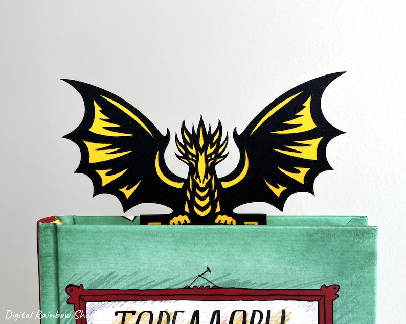 May include: A black and yellow dragon bookmark with outstretched wings, resting on a green book. The dragon's wings have yellow accents. The book has a red framed illustration on the cover.