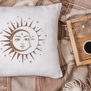 Op de afbeelding: Een wit kussen met een bruin zon- en maandesign. De zon heeft een gezicht en de maan is een halve maan. Het ontwerp is omringd door kleine sterren.