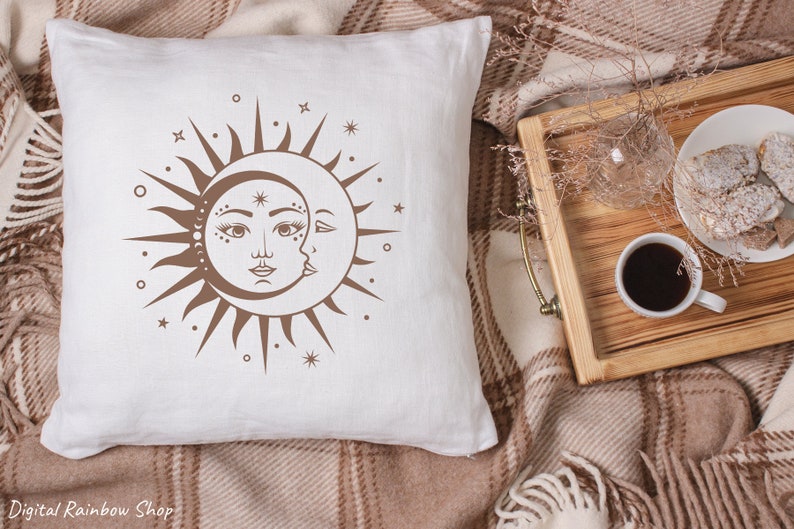 Op de afbeelding: Een wit kussen met een bruin zon- en maandesign. De zon heeft een gezicht en de maan is een halve maan. Het ontwerp is omringd door kleine sterren.