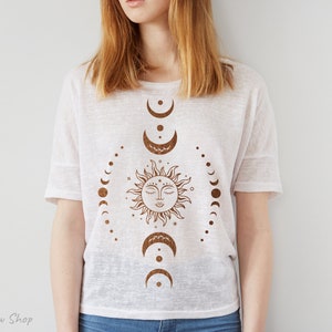 Op de afbeelding: Een wit T-shirt met een bruin zon- en maandesign. De zon heeft een gezicht en de maanfasen zijn in een cirkel eromheen gerangschikt.