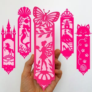 Könnte beinhalten: Eine Sammlung pinkfarbener Papier-Lesezeichen mit verschiedenen ausgeschnittenen Designs. Die Designs umfassen ein Schloss mit einem Einhorn, eine Meerjungfrau, eine Fee mit einem Schmetterling, eine Prinzessin und eine Katze. Der Text "SVG, PNG, DXF, EPS, PDF, JPG" ist ebenfalls enthalten.