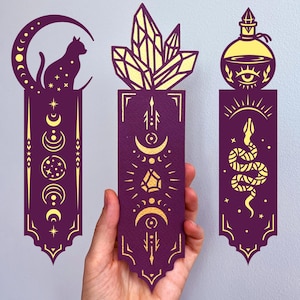 May include: Three purple and gold bookmarks with celestial and mystical designs. One features a cat on a crescent moon, another a crystal, and the last a potion bottle with a snake. The bookmarks are held by a hand.
