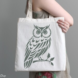 Op de afbeelding: Een witte canvas tote bag met een groen uil ontwerp. De uil is gestileerd met een tribale patroon en zit op een tak.