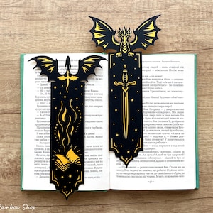May include: Two black and gold dragon-themed bookmarks with detailed cut-out designs. One shows a sword, the other a dragon with wings. They are placed in an open book, creating a fantasy atmosphere.