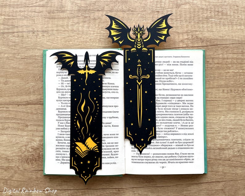 May include: Two black and gold dragon-themed bookmarks with detailed cut-out designs. One shows a sword, the other a dragon with wings. They are placed in an open book, creating a fantasy atmosphere.