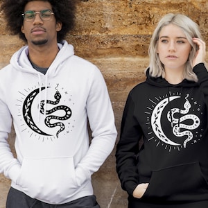 Op de afbeelding: Twee mensen dragen zwarte en witte hoodies met een afbeelding van een slang die om een halve maan gewikkeld is. De afbeelding is in zwart en wit en heeft een hemels thema.