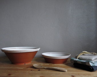 May include: Two terracotta bowls with white rims, one larger than the other, on a wooden surface. A wooden spoon is in front of the bowls. A stack of fabric towels and a dried stalk of wheat are in the background.
